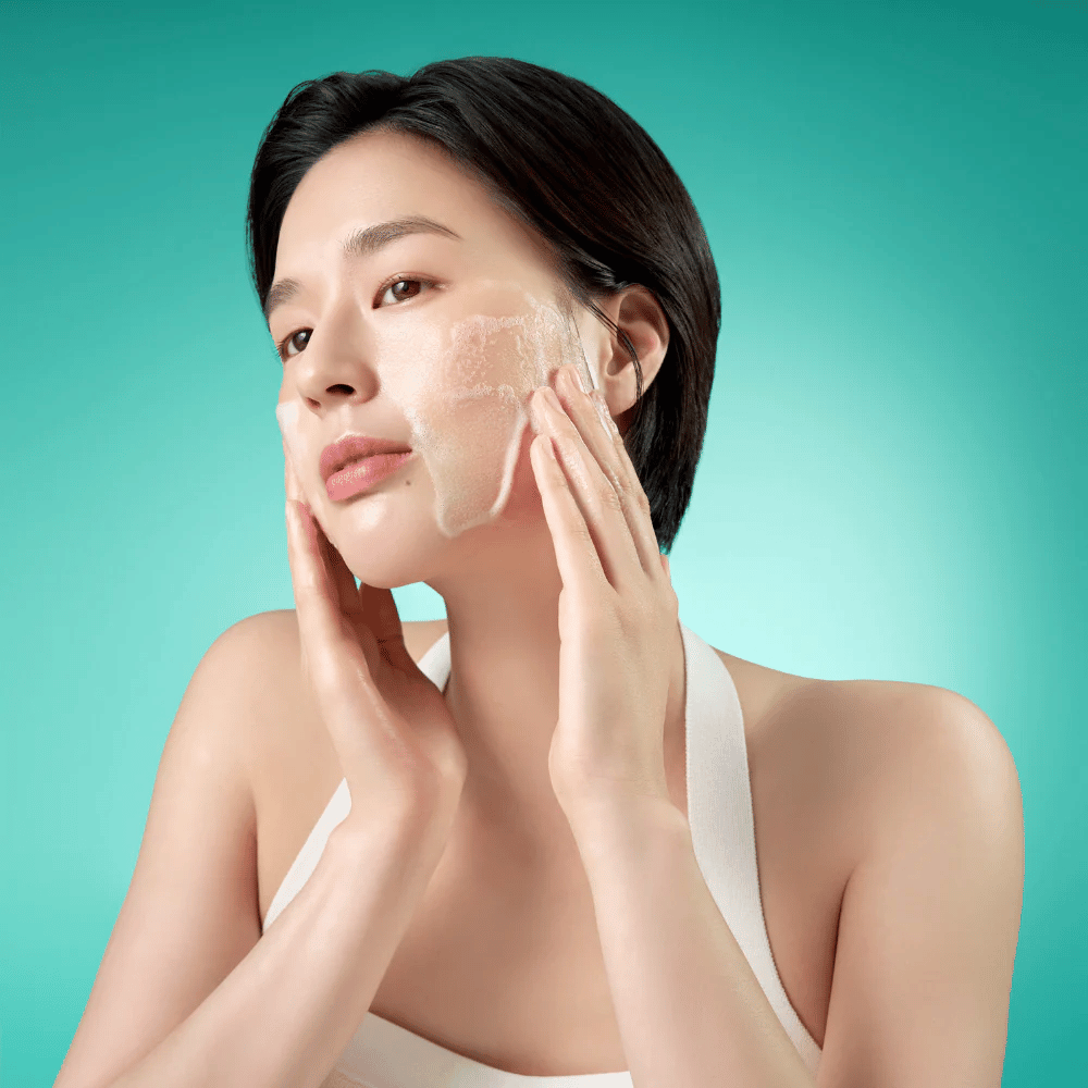 Mujer limpia su rostro con una espuma limpiadora facial suave contra un fondo verde.