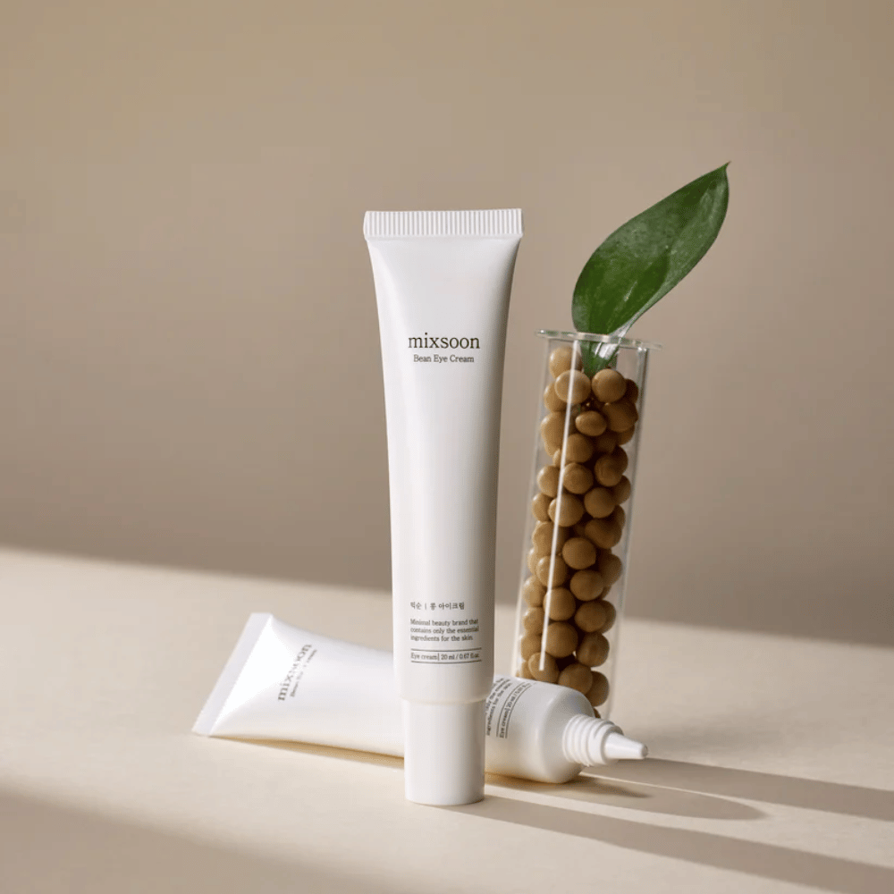 Dos tubos Mixsoon Bean Eye Cream colocados junto a un tubo de ensayo lleno de semillas de soja y una hoja verde, sobre una superficie beige con juegos de sombras.