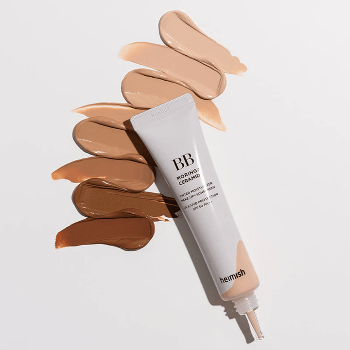 Heimish Moringa Ceramide BB Cream rodeado de varias muestras de tono en diferentes tonos de piel. La imagen muestra la amplia gama de colores del producto y la textura cremosa que proporciona una cobertura natural y un tono de piel uniforme.