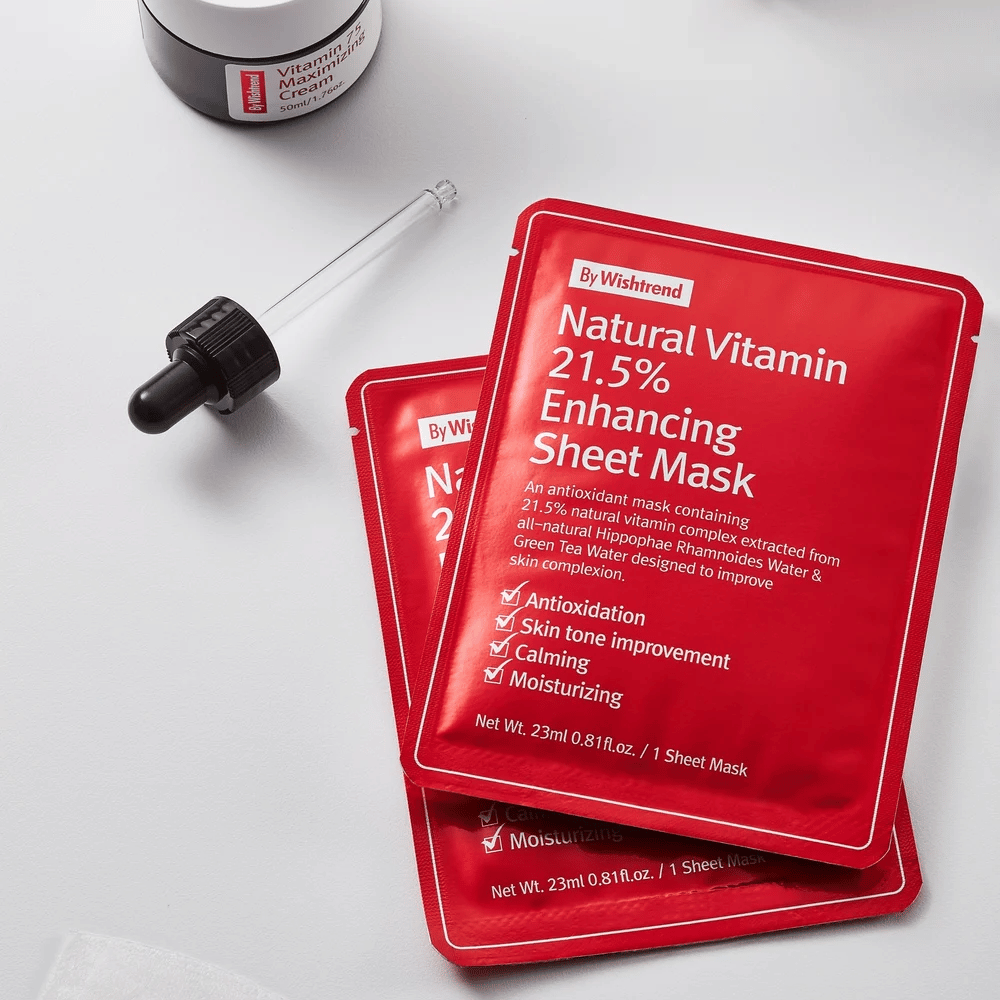 By Wishtrend Natural Vitamin 21.5% Enhancing Sheet mask en envases rojos, colocados sobre una superficie blanca junto con una pipeta y un tarro de crema facial.