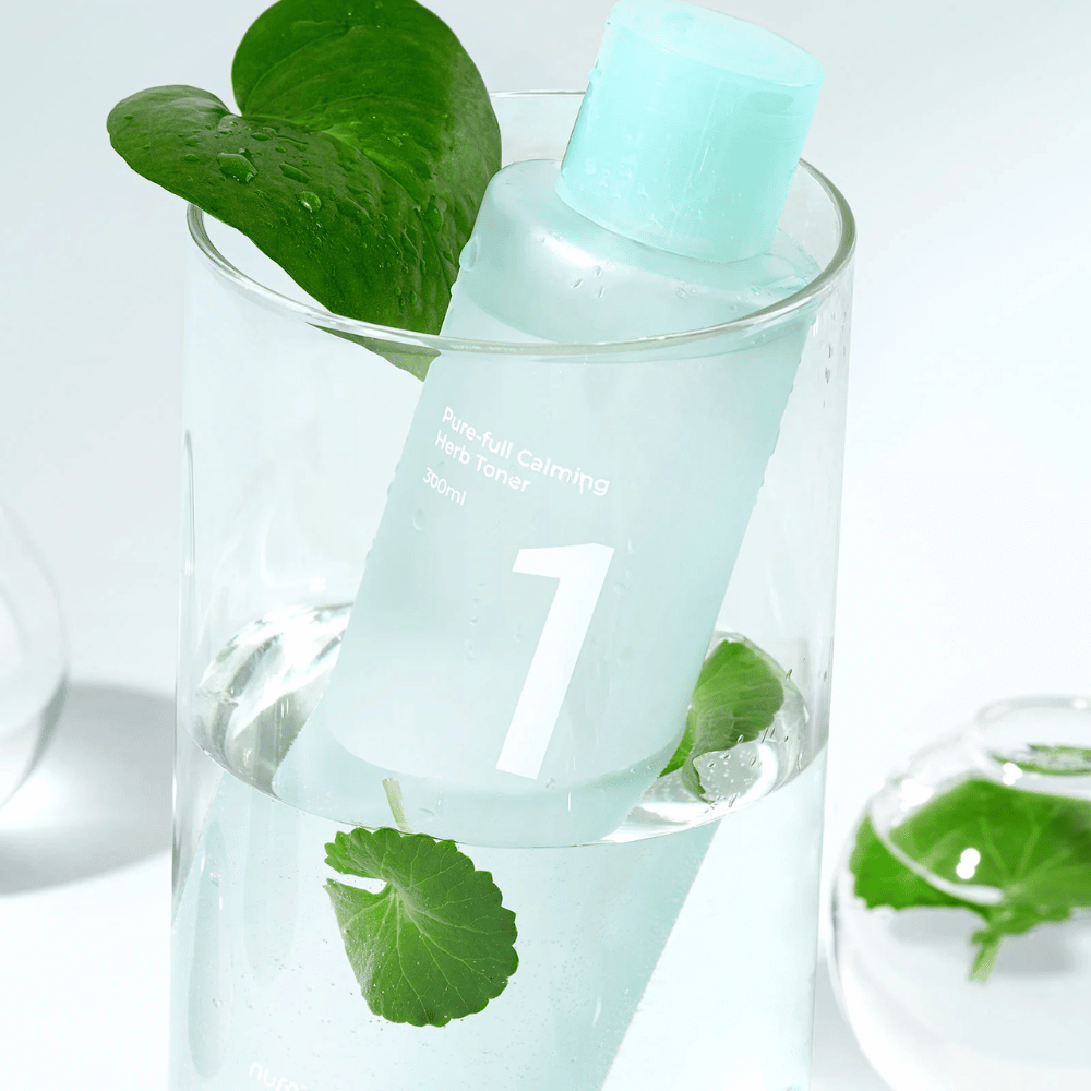 Una botella con No.1 Pure-Full Calming Herb Toner colocada en un vaso con agua y hojas verdes. La botella tiene una tapa azul claro y una etiqueta con el texto "Pure-full Calming Herb Toner" y "300ml". El producto se ve fresco con agua y vegetación en el fondo, lo que da una sensación calmante y natural.