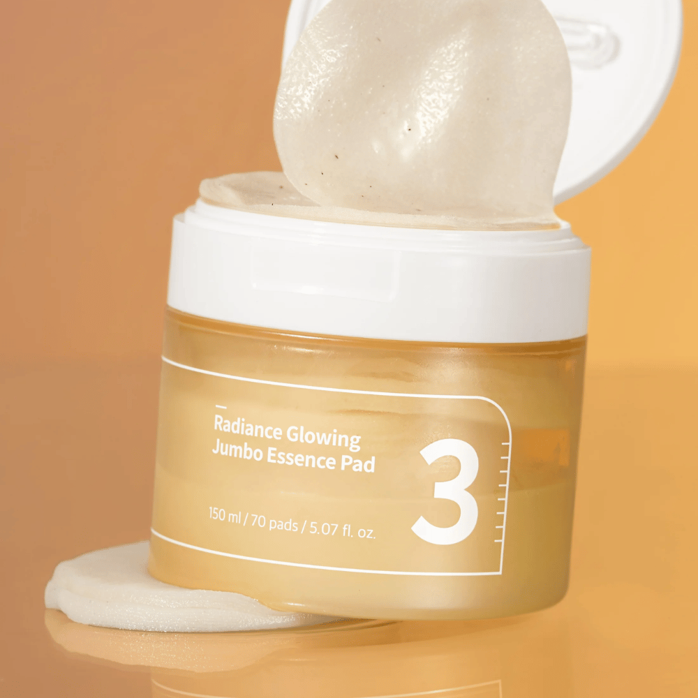 Frasco abierto con numbuzin No. 3 Radiance Glowing Jumbo Essence Pad, una almohadilla se ve inclinada contra el borde, fondo en tono naranja.