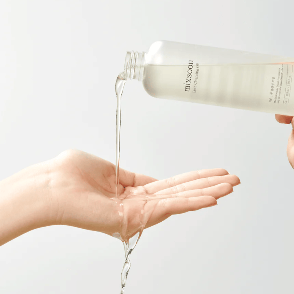Un aceite limpiador claro se vierte de una botella transparente con una etiqueta blanca donde dice "MixSoon" en una palma abierta. El aceite forma un chorro brillante entre la apertura de la botella y la mano, demostrando la consistencia líquida del aceite. La mano está quieta y se sostiene de manera que pueda recoger toda la cantidad de aceite limpiador contra un fondo claro de un solo color.