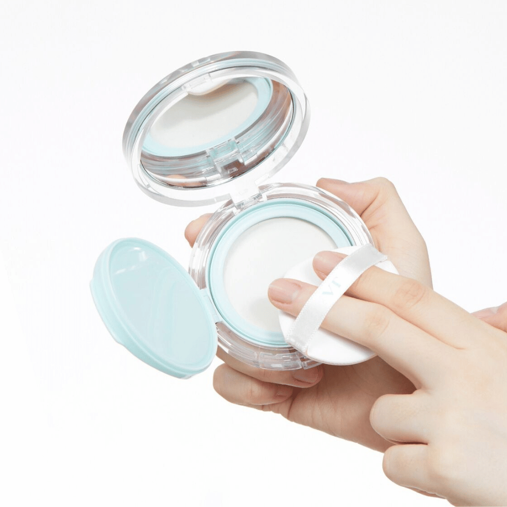 Una mano sostiene un tarro abierto con VT PDRN Essence Glow Sun Pact. La borla está a punto de aplicar el polvo, que tiene una superficie blanca y un borde azul alrededor del tarro.