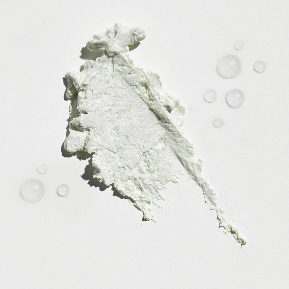 Primer plano de una sustancia cremosa blanca sobre un fondo blanco, con algunas gotas de agua alrededor. La textura parece ser un polvo solar o foundation con un ligero brillo.