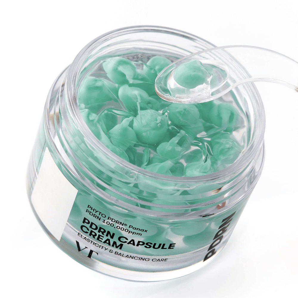 Primer plano de un frasco abierto con VT PDRN Capsule Cream. El contenido consiste en pequeñas cápsulas verdes en un gel transparente, con una espátula incluida para la aplicación. El texto en el frasco dice "Elasticity & Balancing Care".