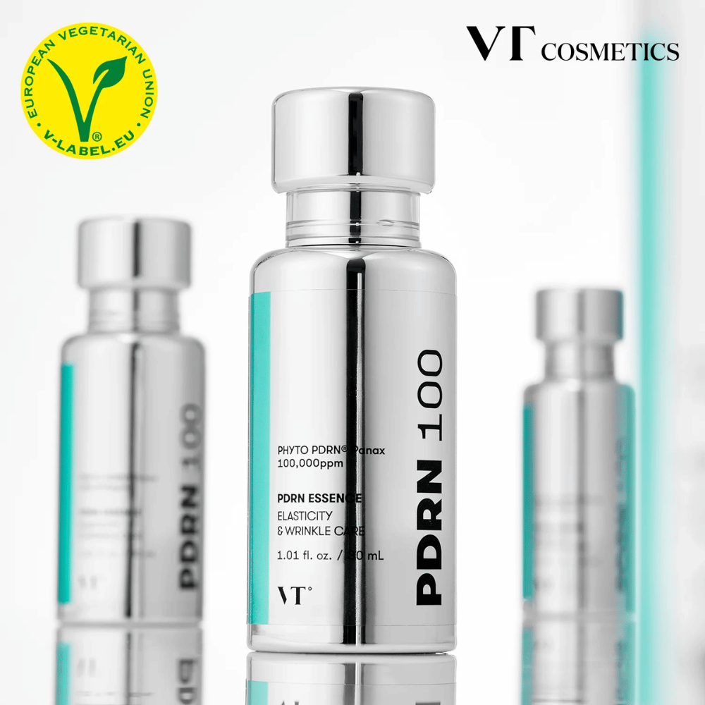 Varias botellas plateadas de VT PDRN 100 Essence están alineadas, con una clara indicación de 100,000 ppm PDRN y un enfoque en elasticidad y cuidado de arrugas. En la parte superior se ve el logotipo de VT Cosmetics y una etiqueta V para contenido vegetariano.