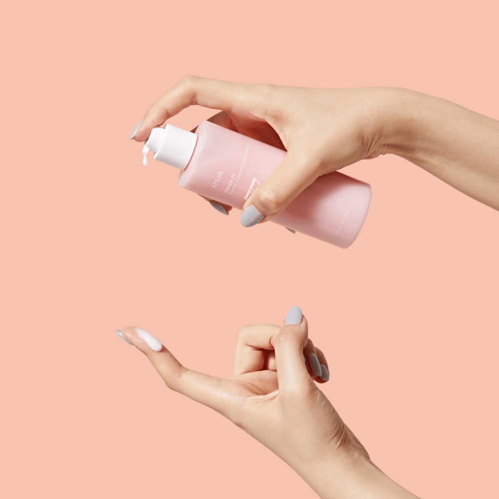 La imagen muestra las manos de alguien sosteniendo una botella con ANUA Peach 77% Niacin Conditioning Milk. Este producto para el cuidado de la piel parece tener una consistencia lechosa y está diseñado para cuidar y mejorar el estado de la piel.