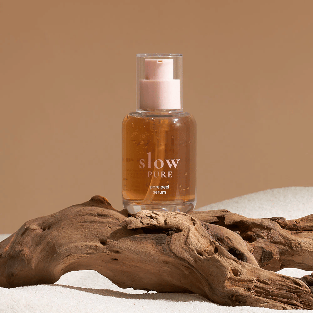 Slow Pure Pore Peel Serum colocada sobre arena con detalle de madera en el fondo, símbolo de cuidado de la piel natural.