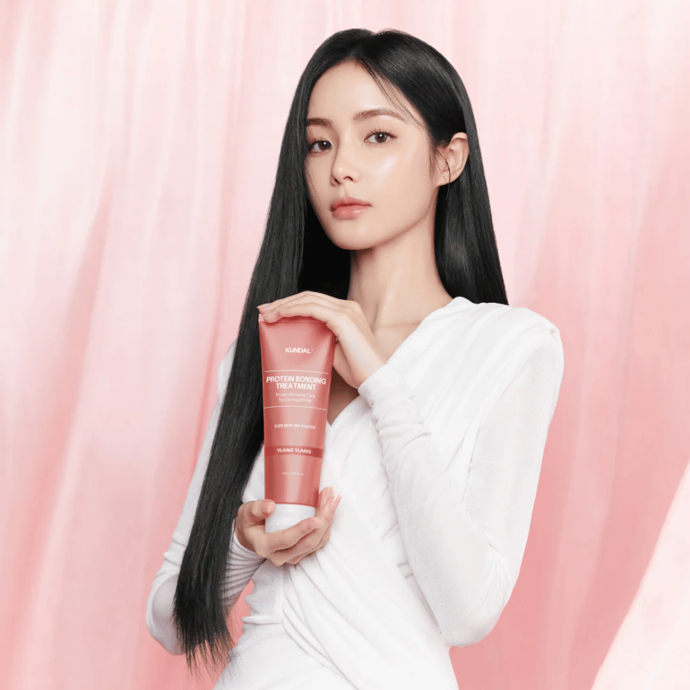 Mujer con cabello largo y brillante sostiene KUNDAL Protein Bonding Treatment Violet Muguet contra un fondo rosa.