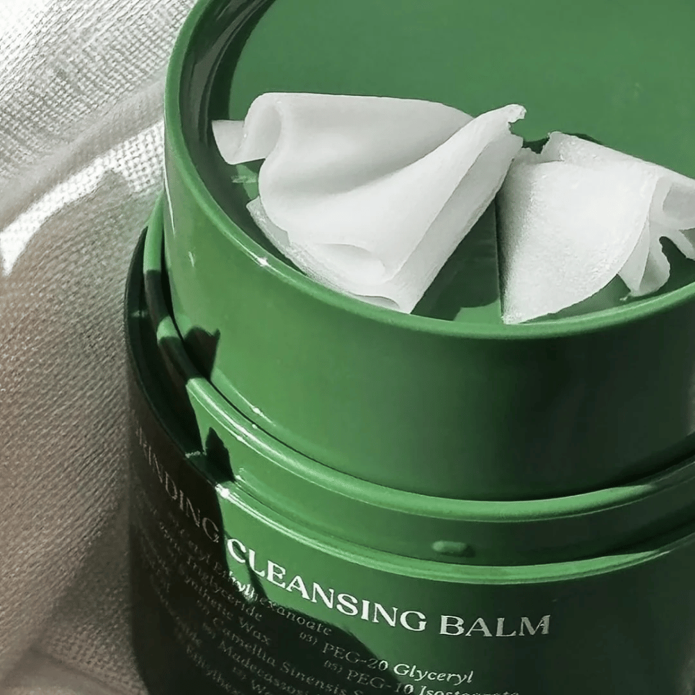 La imagen muestra un tarro verde con "Cleansing Balm", que está abierto para revelar la espátula incluida y el contenido suave. La consistencia cremosa refleja la luz, lo que da una sensación de lujo y eficacia en la limpieza.