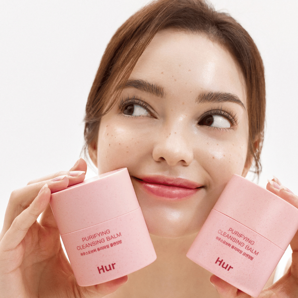Modelo sostiene dos tarros Cómo Purifying Cleansing Balm. Bálsamo limpiador suave que es adecuado para piel sensible y deja una sensación fresca e hidratada después de la limpieza.