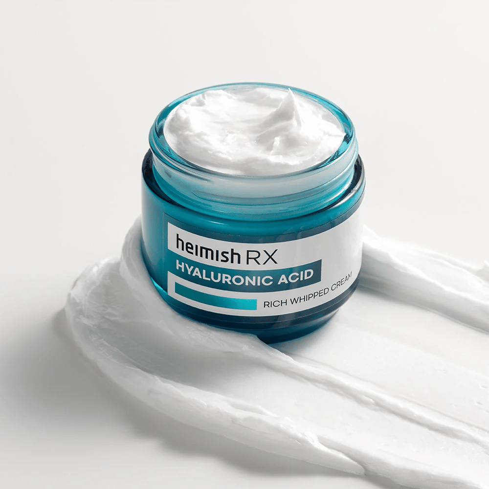 Heimish RX Hyaluronic Acid Rich Whipped Cream en un tarro de vidrio azul con consistencia rica y esponjosa, esparcida sobre un fondo blanco. Crema facial intensamente hidratante con ácido hialurónico para una hidratación profunda y piel suave.