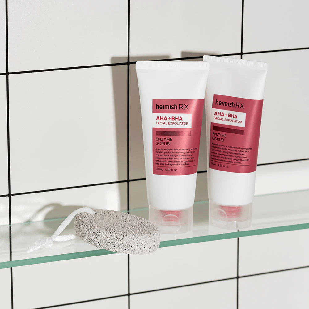 Dos tubos Heimish RX AHA + BHA Facial Exfoliator colocados en un estante de vidrio en un baño con pared de azulejos blancos. Al lado de los productos hay una piedra pómez con una cuerda blanca. Una imagen de cuidado de la piel elegante y minimalista.