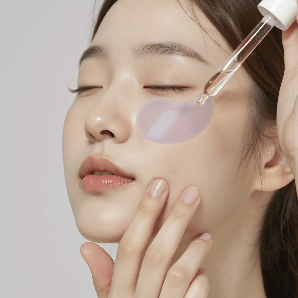 Una mujer con un parche de hidrogel morado para los ojos aplica suero debajo del ojo con una pipeta. El producto hidrata y cuida la piel sensible alrededor de los ojos.