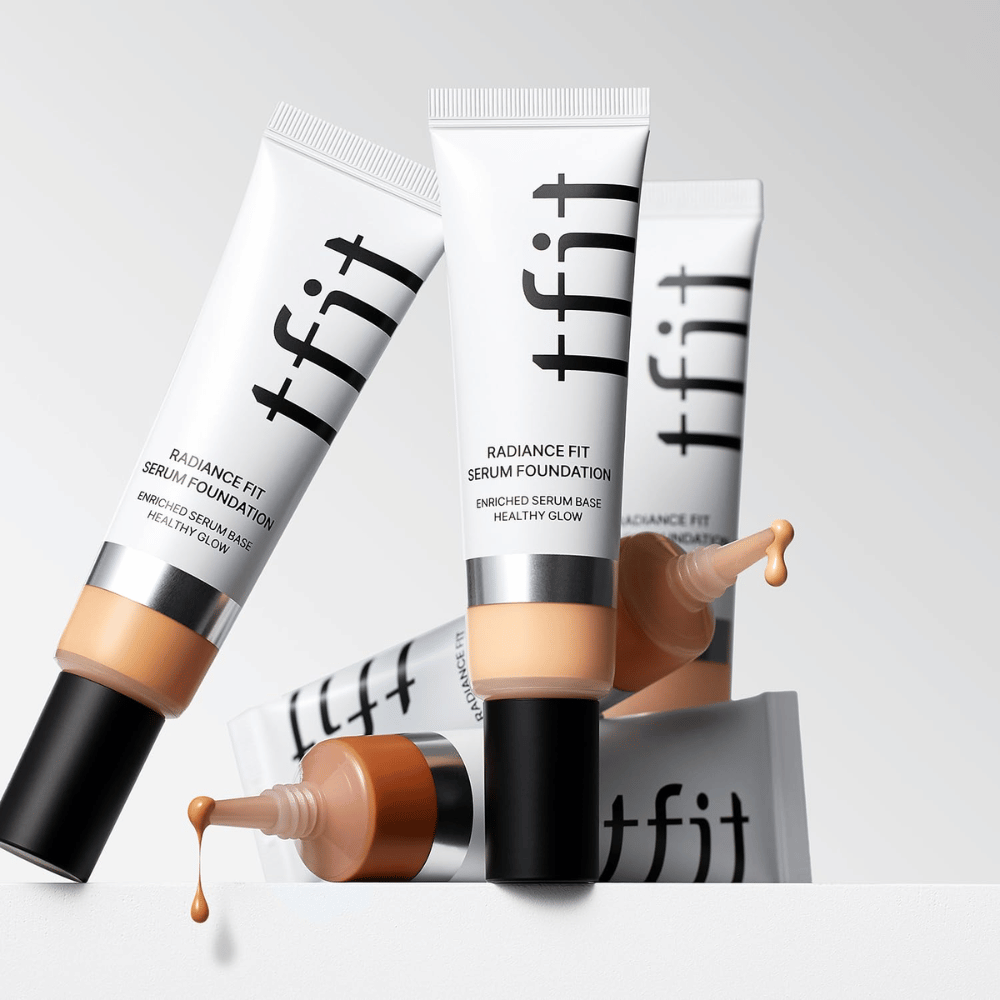Varios tubos de TFIT Radiance Fit Serum Foundation en diferentes tonos, uno de los cuales tiene foundation goteando, sobre un fondo gris claro.