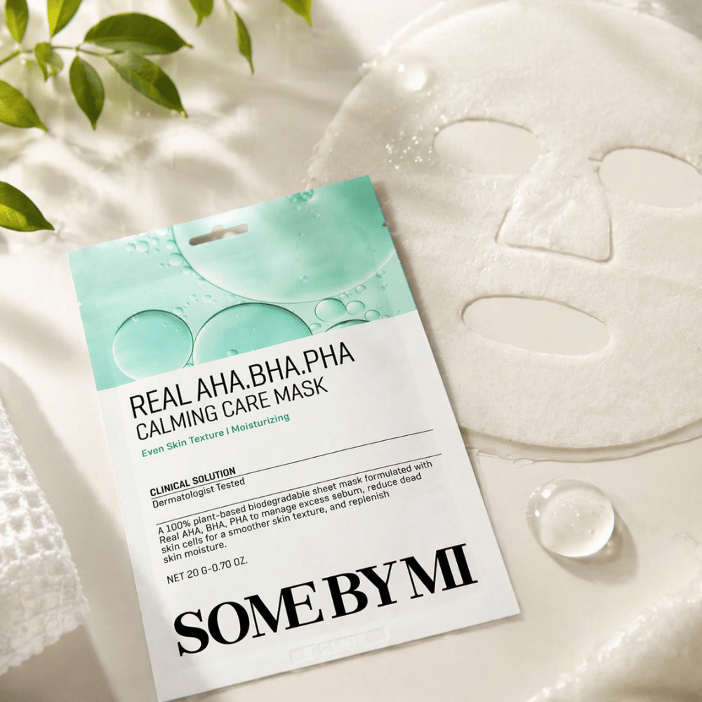 SOME BY MI Real AHA.BHA.PHA Calming Care Mask se muestra junto con la mascarilla de tela desplegada. Alrededor del producto se ven gotas de humedad y hojas verdes que refuerzan la sensación de hidratación, cuidado calmante y exfoliación suave. La imagen destaca el enfoque de la mascarilla en una textura de la piel más uniforme y un equilibrio de hidratación mejorado.