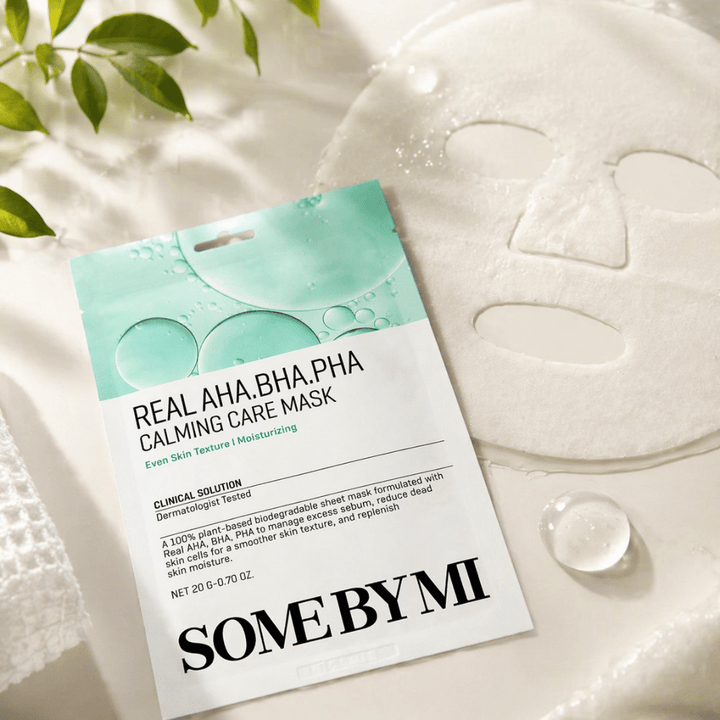SOME BY MI Real AHA.BHA.PHA Calming Care Mask se muestra junto con la mascarilla de tela desplegada. Alrededor del producto se ven gotas de humedad y hojas verdes que refuerzan la sensación de hidratación, cuidado calmante y exfoliación suave. La imagen destaca el enfoque de la mascarilla en una textura de la piel más uniforme y un equilibrio de hidratación mejorado.