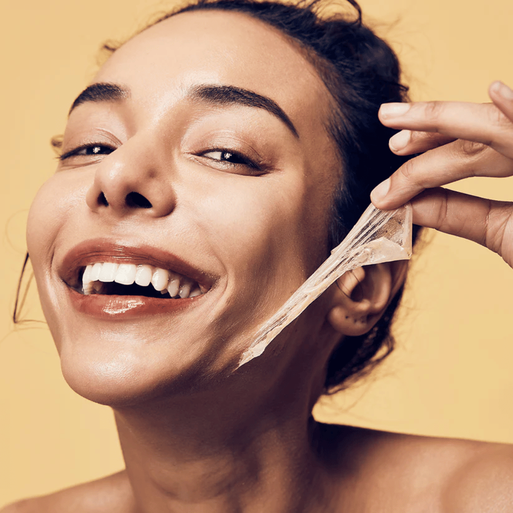 La imagen muestra a una mujer con una gran sonrisa mientras se quita una mascarilla peel-off de su rostro. La mujer parece feliz y satisfecha mientras usa el producto de belleza, lo que transmite una sensación de bienestar y cuidado de la piel.