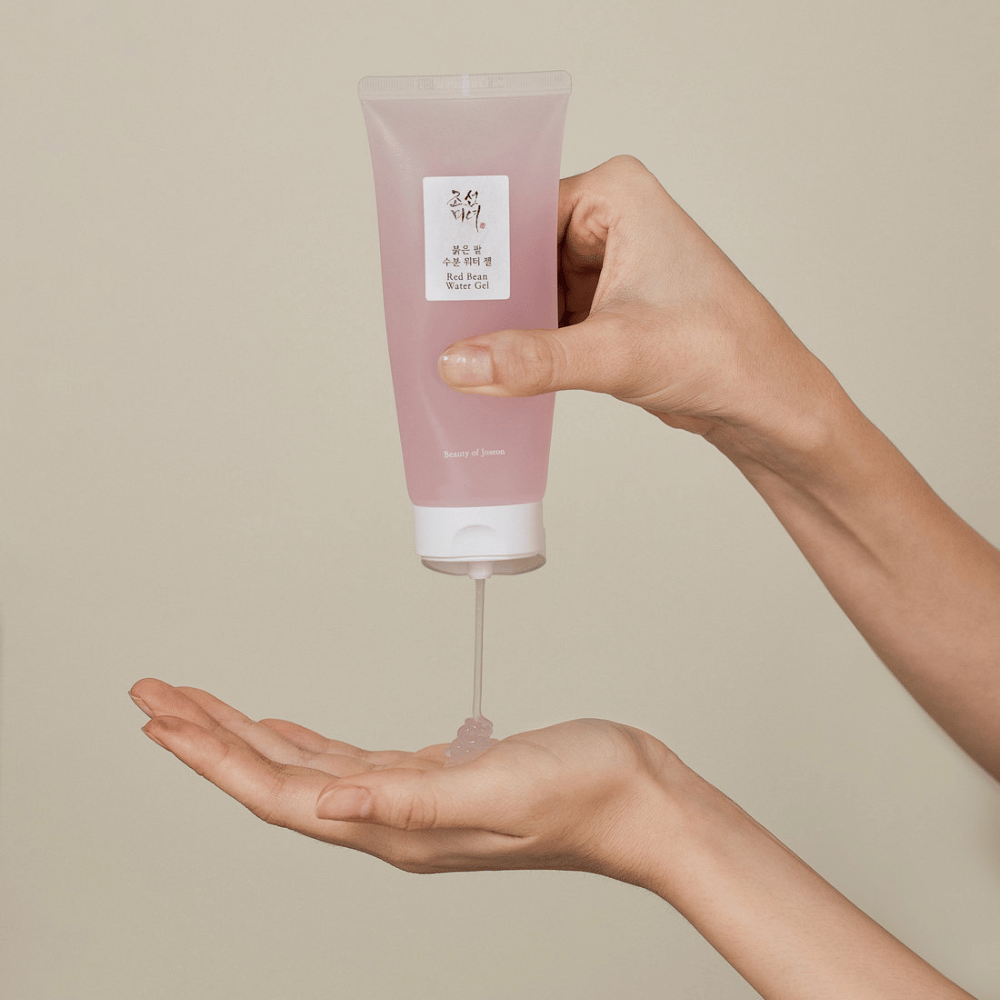 La imagen muestra a una persona sosteniendo un tubo de "Red Bean Water Gel" de Beauty of Joseon y exprimiendo un gel transparente en su mano. El producto parece estar destinado al cuidado de la piel y tiene un empaque atractivo y suave de color rosa.