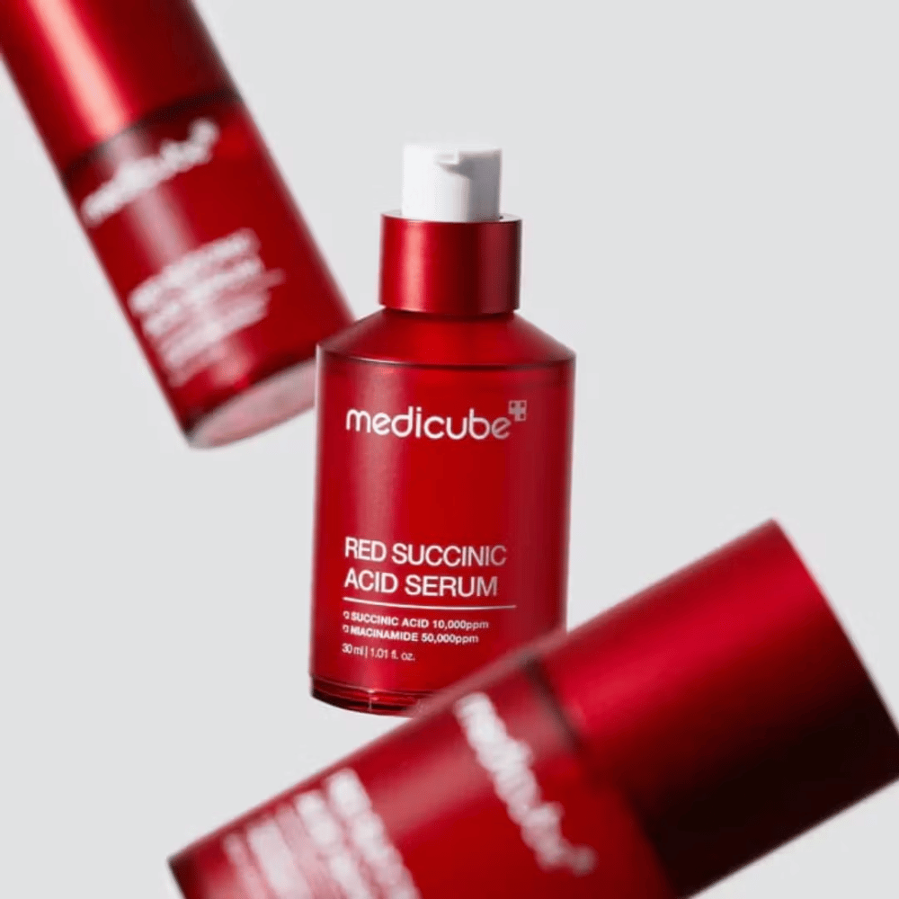 Medicube Red Succinic Acid Serum en botella roja con bomba, rodeada de más botellas idénticas contra un fondo claro. El producto contiene ácido succínico y niacinamida para tratar el acné y las manchas de pigmentación.