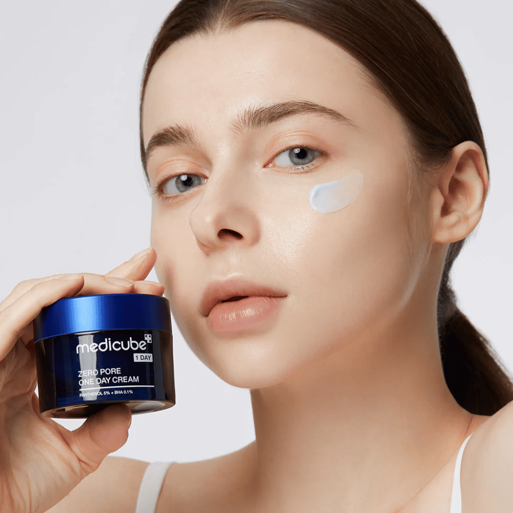 Mujer con un look natural sostiene Medicube Zero Pore One Day Cream. Se ha aplicado una pequeña cantidad de crema en su mejilla. El producto está diseñado para mejorar la textura de la piel y minimizar el tamaño de los poros, con una fórmula ligera e hidratante.