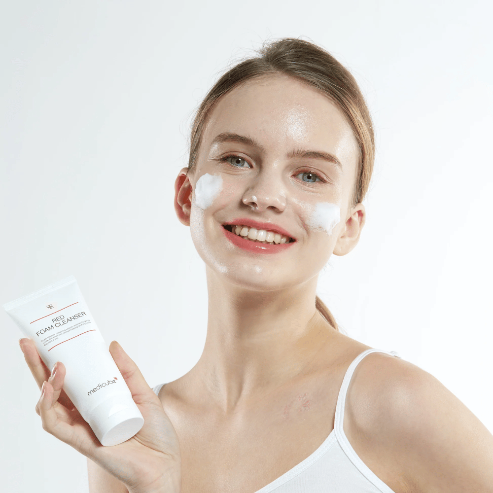 Joven con espuma limpiadora en el rostro sostiene un tubo de Medicube Red Foam Cleanser y sonríe a la cámara. Lleva una camiseta blanca y el fondo es claro y sencillo.