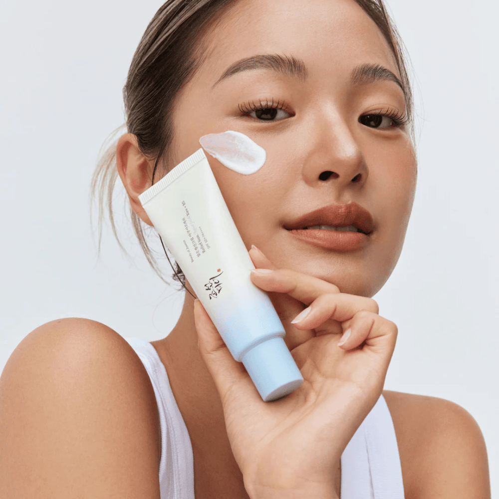 Mujer sostiene Beauty of Joseon Relief Sun: crema solar Aqua-Fresh Rice + B5 y muestra su consistencia cremosa en la mejilla. Un producto solar ligero que hidrata y protege la piel con SPF 50+ PA++++.
