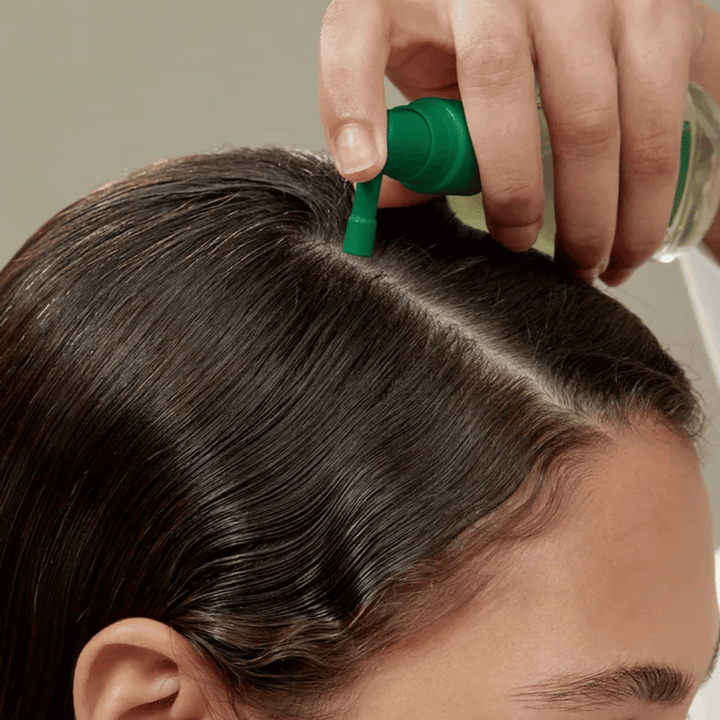 Una persona aplica AROMATICA Rosemary Root Enhancer directamente en el cuero cabelludo con ayuda de la boquilla de precisión. La imagen muestra cómo se utiliza el producto para el cuidado del cuero cabelludo y el fortalecimiento de la raíz del cabello.