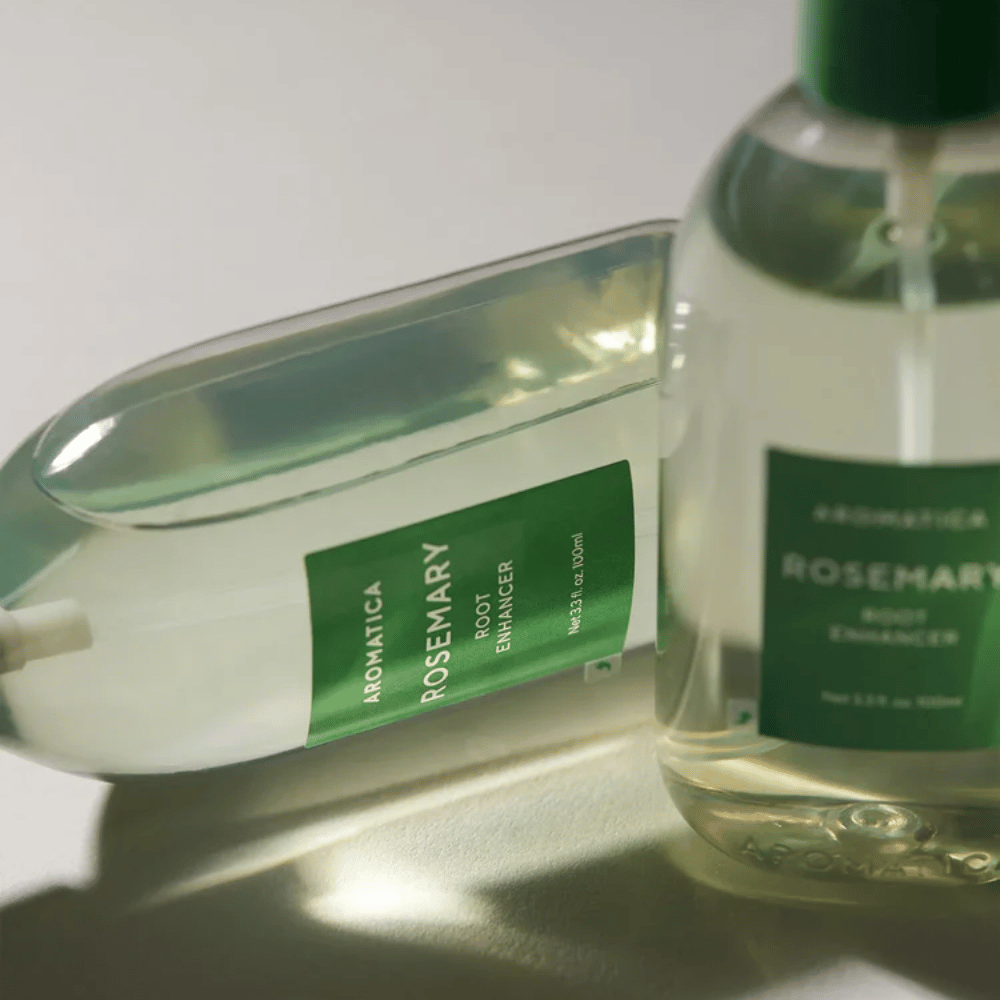 Dos botellas de AROMATICA Rosemary Root Enhancer con tapa protectora transparente y etiqueta verde. Primer plano que resalta el diseño premium del producto y su concepto natural para el cuidado capilar.