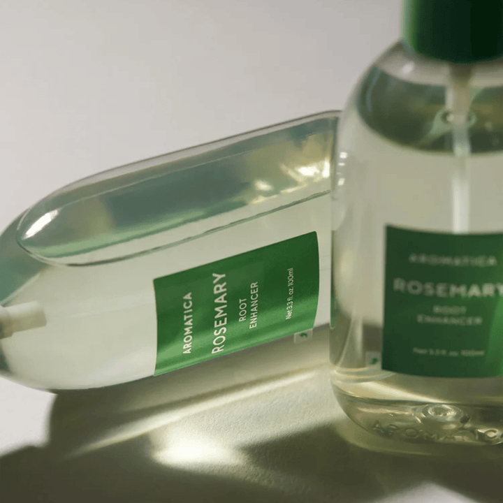 Dos botellas de AROMATICA Rosemary Root Enhancer con tapa protectora transparente y etiqueta verde. Primer plano que resalta el diseño premium del producto y su concepto natural para el cuidado capilar.