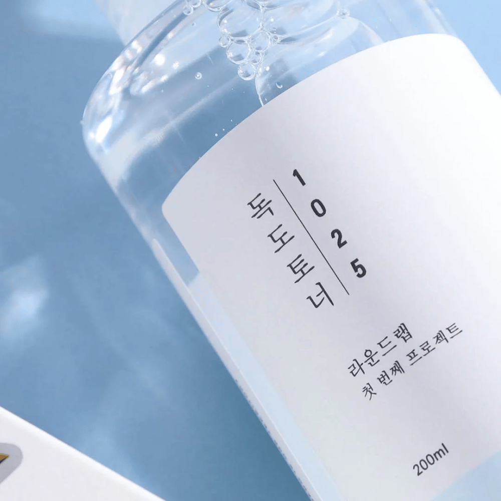 Primer plano de una botella transparente Round Lab 1025 Dokdo Toner, llena de líquido claro, contra un fondo azul.