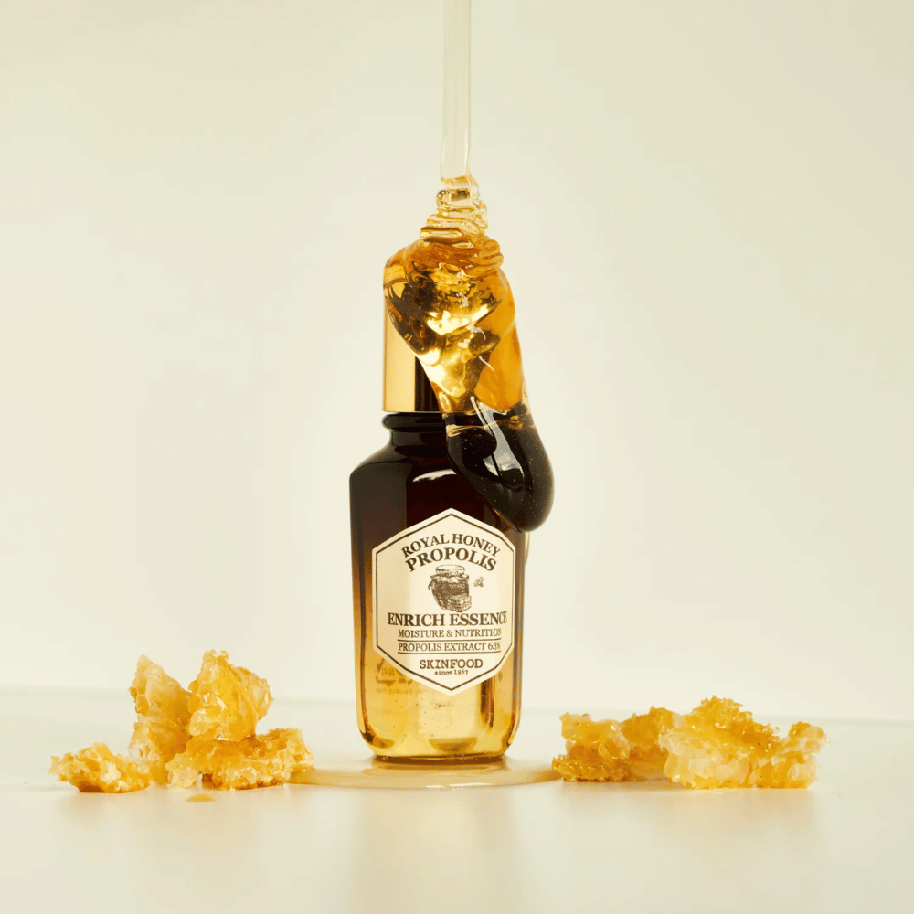 Imagen de una botella de vidrio con Royal Honey Propolis Enrich Essence de Skinfood. Una gran gota de miel dorada cae desde la tapa dorada de la botella. Alrededor de la botella hay pequeños trozos de panal esparcidos sobre una superficie clara. La esencia se comercializa con un enfoque en la humedad y la nutrición, con un 63% de extracto de propóleo.