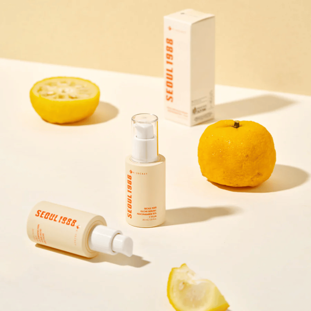 Varias botellas y empaques de Seúl 1988 Glow Serum dispuestos con frutas de yuja enteras y partidas por la mitad en el fondo.