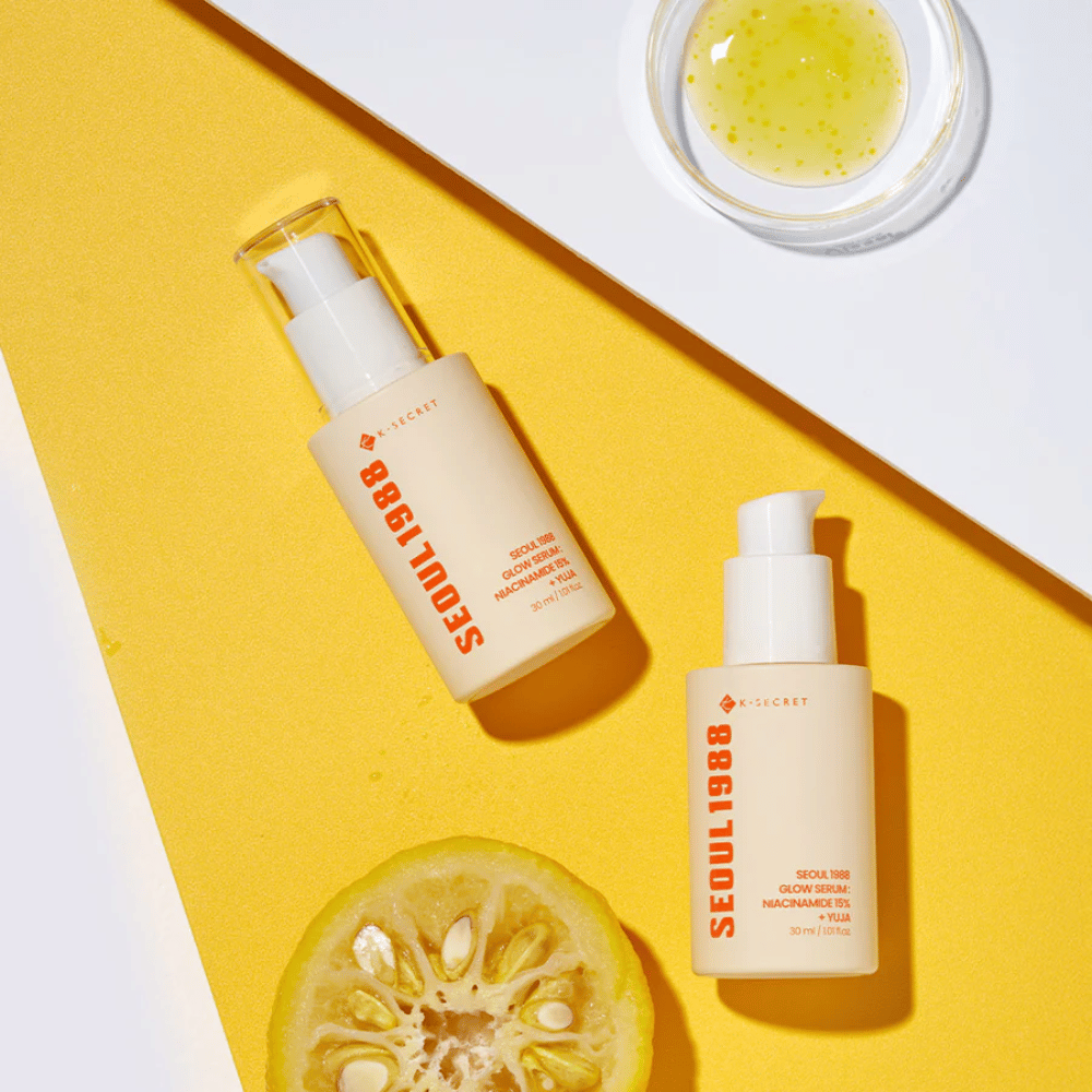 Dos botellas de Seúl 1988 Glow Serum colocadas sobre un fondo amarillo junto con un cuenco con suero amarillo y una fruta cítrica partida por la mitad.