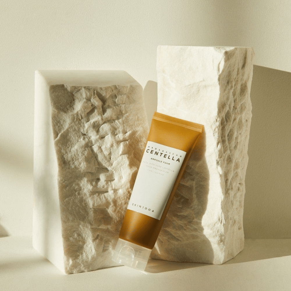 SKIN1004 Madagascar Centella Ampoule Foam de pie contra bloques de piedra claros bajo luz solar natural, con su diseño de tubo marrón y blanco y minimalismo elegante.