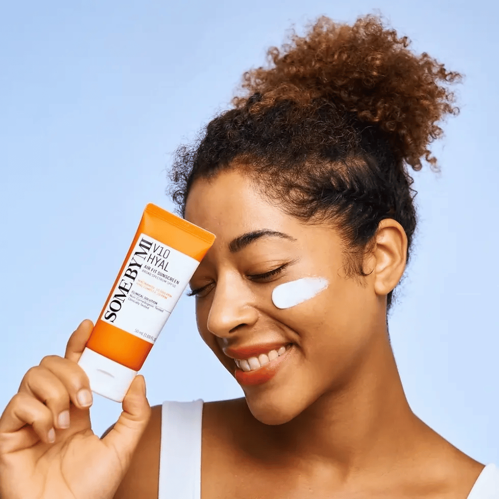Una mujer sostiene un tubo de SOME BY MI V10 Hyal Air Fit Sunscreen y tiene una mancha blanca de protector solar en la mejilla. Ella sonríe alegremente y muestra claramente el producto, destacando su uso y beneficios para el cuidado de la piel y la protección solar.