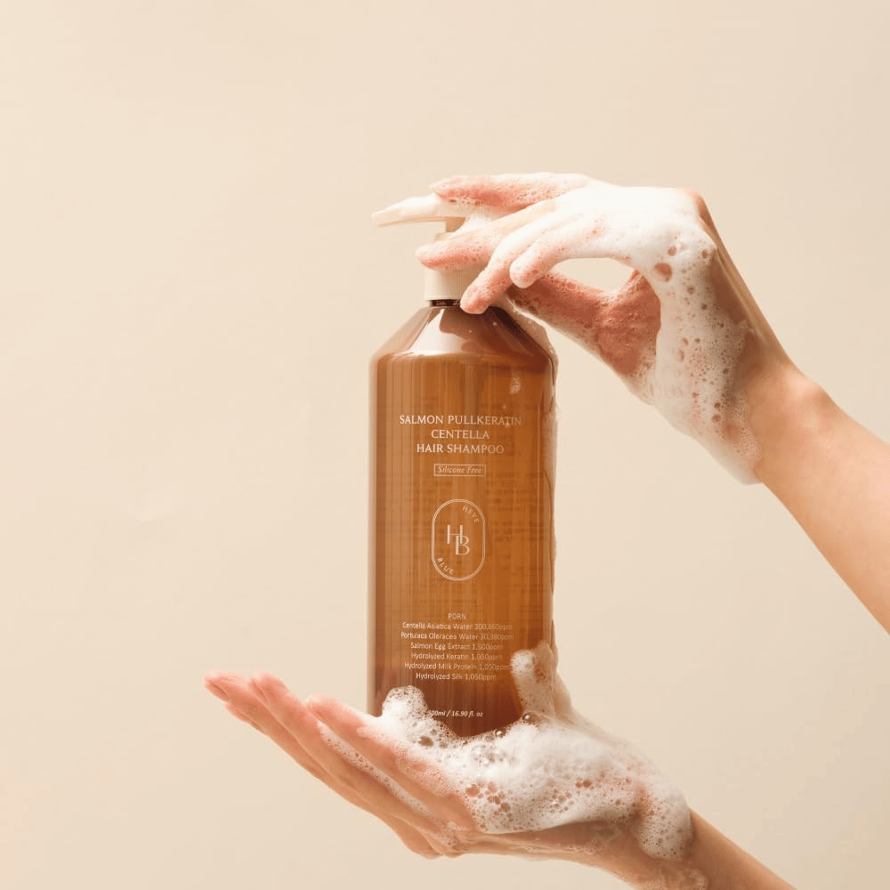 Heveblue Salmon Pullkeratin Centella Hair Shampoo en una botella marrón rodeada de espuma. El producto se sostiene entre dos manos y destaca una experiencia de cuidado capilar limpiadora y nutritiva con abundante espuma.