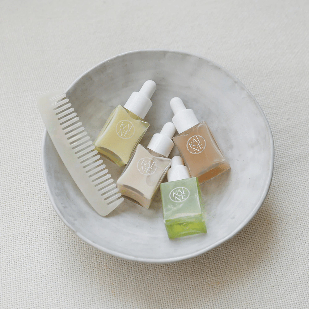 Cuatro pequeños frascos de suero de KAINÉ Serum Discovery Kit están en un cuenco blanco al lado de un peine, con un fondo natural y tranquilo en tono beige.