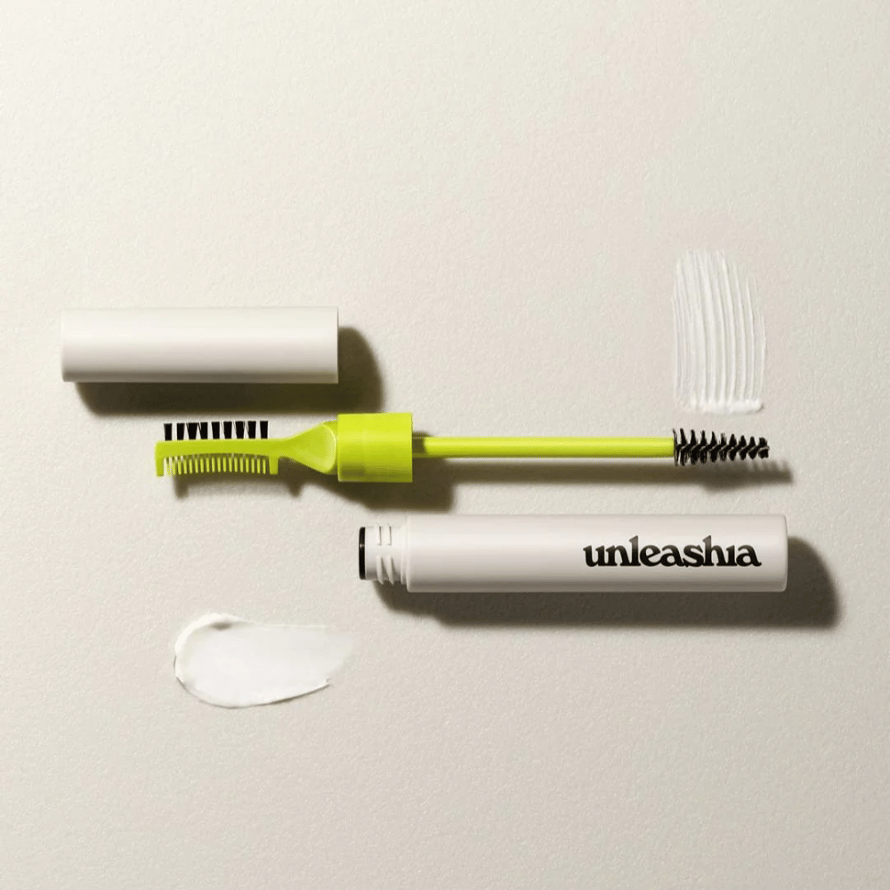 Un set para esculpir cejas con un peine verde, cepillo negro y un tubo blanco etiquetado "unleashia" sobre un fondo claro con un montón de producto transparente.