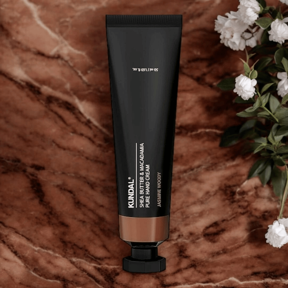 KUNDAL Shea Butter & Macadamia Pure Hand Cream Jasmine Woody colocada sobre mármol con flores – crema de manos nutritiva que cuida manos secas y ofrece un elegante aroma de jazmín amaderado.