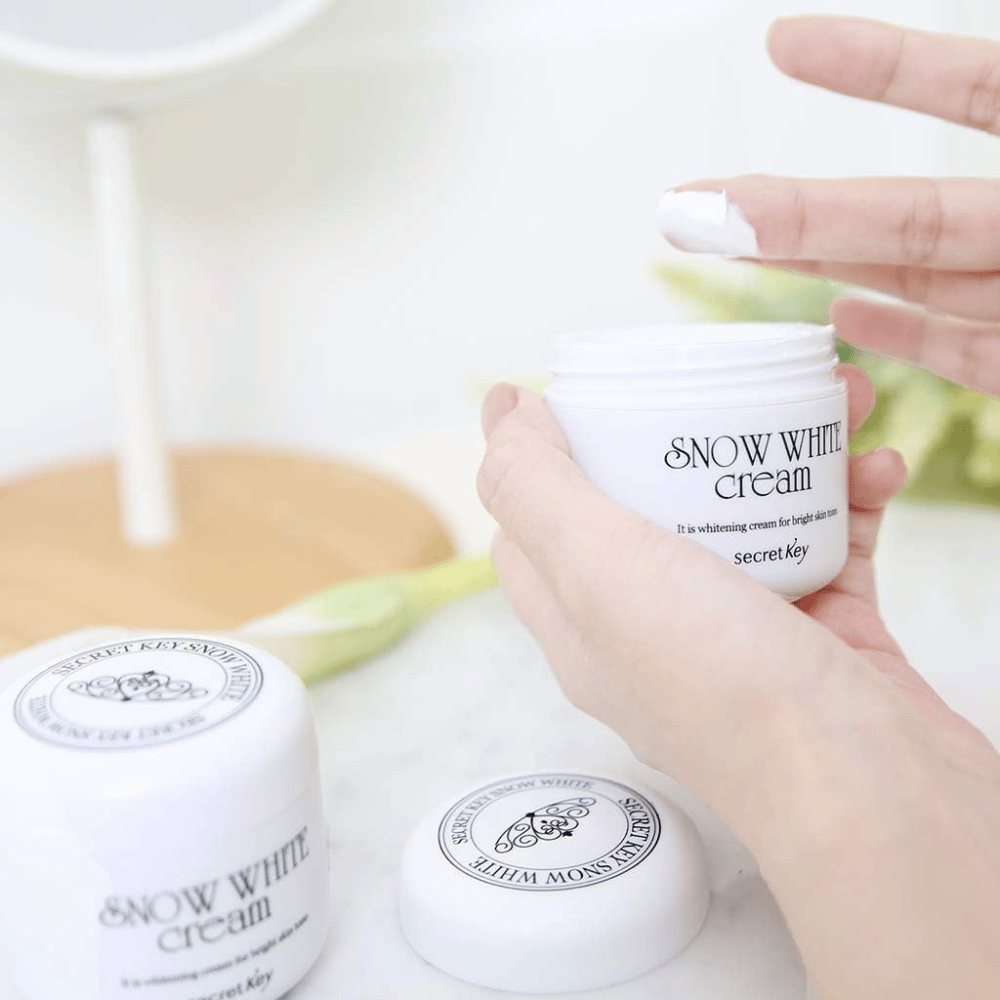 Mano que toma crema blanca de un frasco Secret Key Snow White Cream en un tocador.