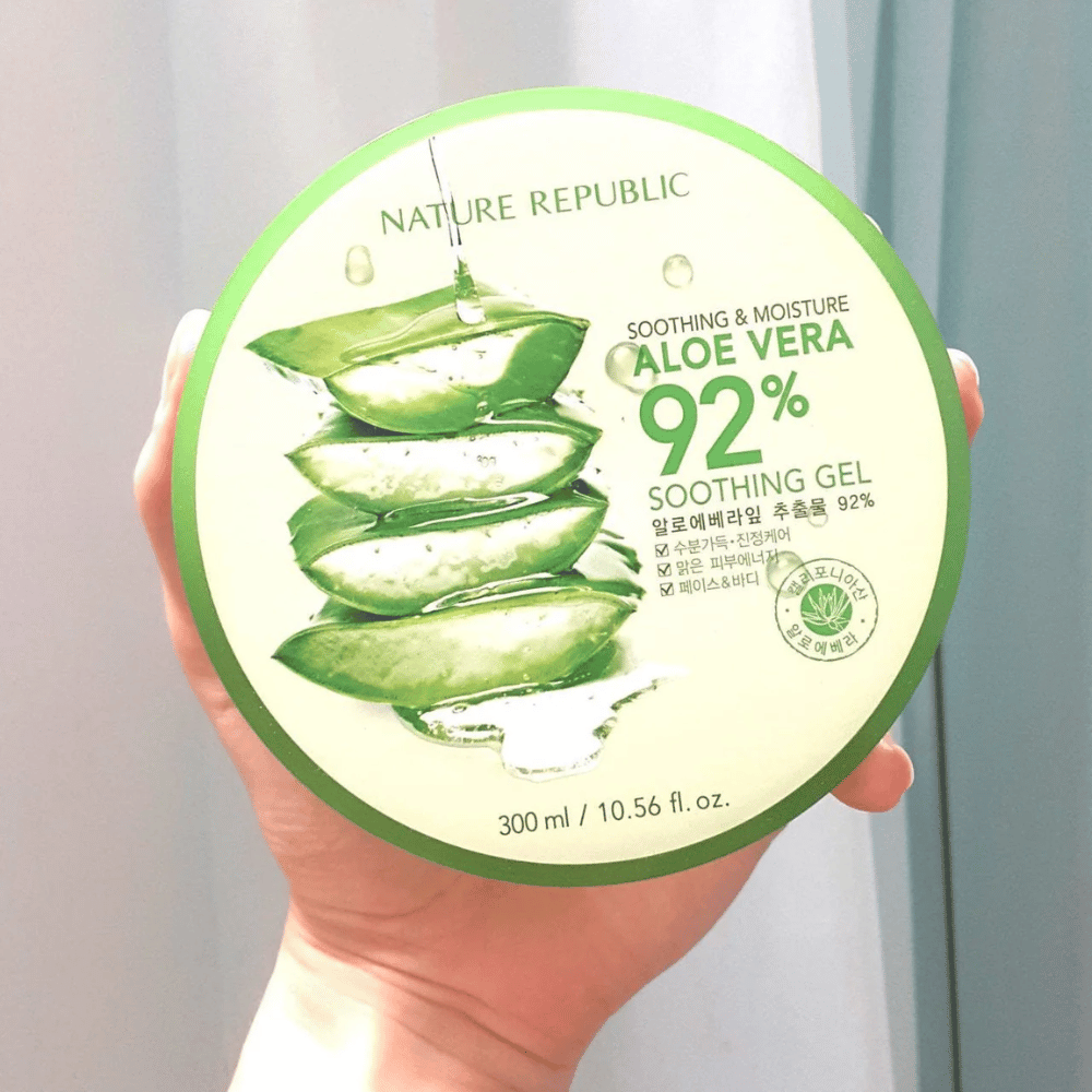 Una mano sostiene un tarro redondo con Nature Republic Aloe Vera 92% Gel Calmante. El tarro es verde con una imagen de aloe vera y texto que describe sus propiedades calmantes e hidratantes. El contenido es de 300 ml.