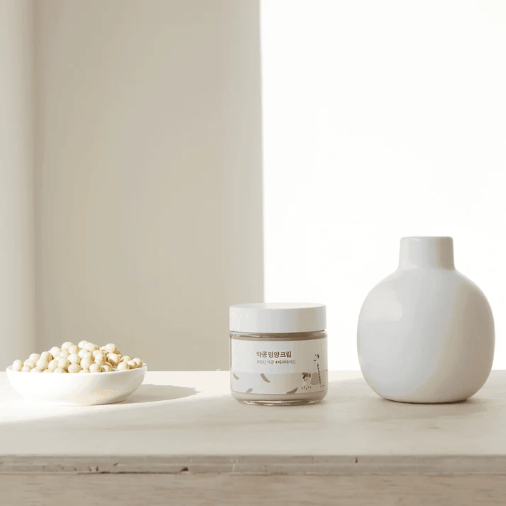 Mixsoon Soybean Nourishing Cream está sobre una mesa de madera junto a un jarrón y soja. Una crema minimalista e hidratante con ingredientes naturales.