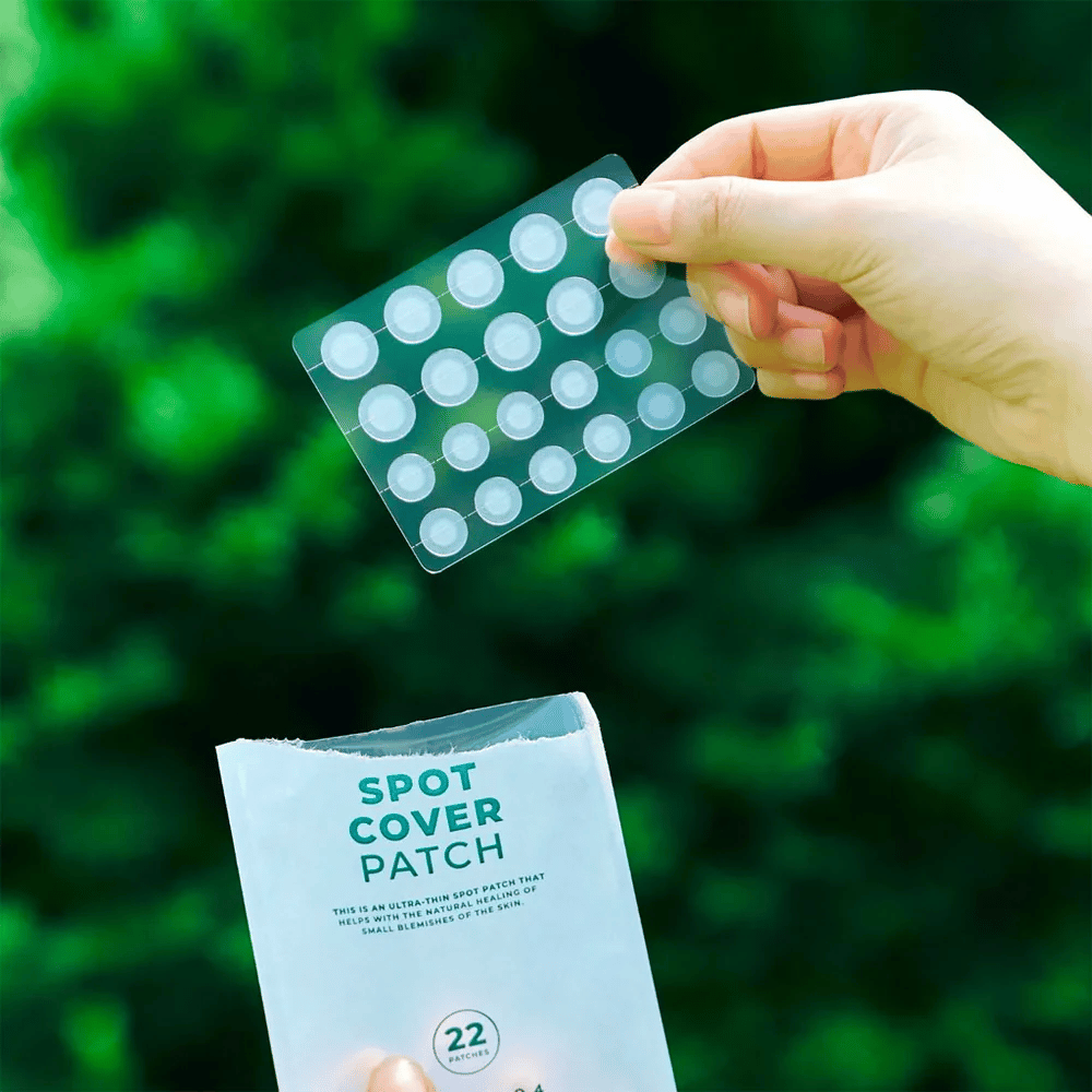 Una mano sostiene una hoja con Spot Cover Patches, que son transparentes y redondeados. En el fondo se ve un entorno verde borroso. El empaque muestra una parte del empaque con el texto "SPOT COVER PATCH" y la información de que el producto contiene 22 parches que ayudan a curar pequeñas áreas de la piel.