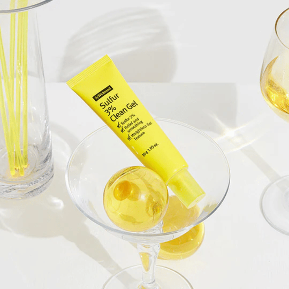 By Wishtrend Sulfur 3% Clean Gel en un tubo amarillo colocado en un vaso de cóctel con una esfera amarilla, rodeado de elegantes copas sobre un fondo blanco.