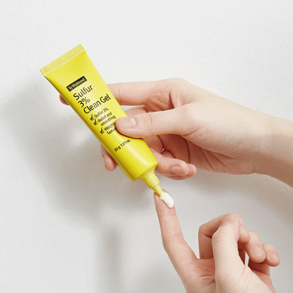 Primer plano de manos aplicando By Wishtrend Sulfur 3% Clean Gel, un tubo amarillo con gel blanco, sobre un fondo blanco.