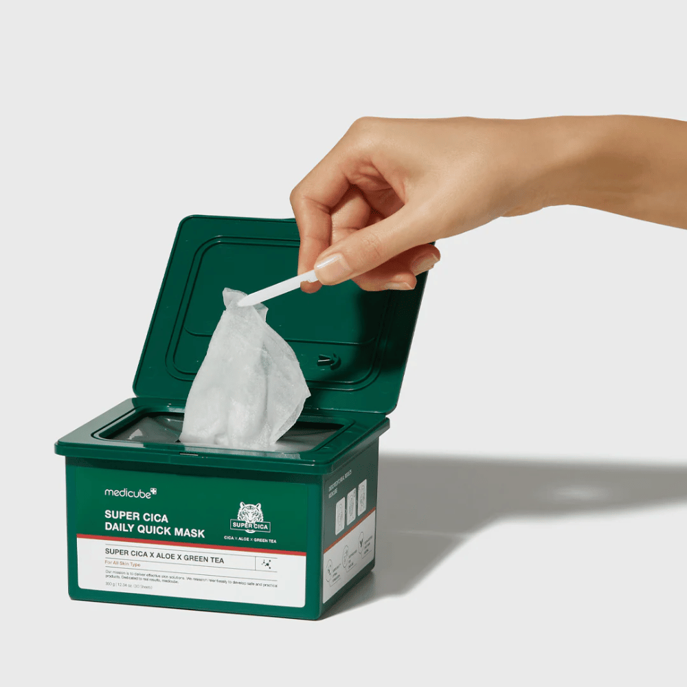 Contenedor verde con tapa abierta muestra medicube Super Cica Daily Quick Mask. Una mano saca una mascarilla de hoja con las pinzas incluidas. El producto contiene Cica, aloe y té verde, ideal para hidratación rápida y efecto calmante.