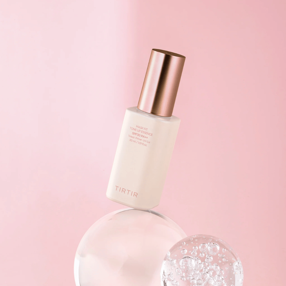 La imagen muestra una botella de "TIRTIR MASK FIT TONE UP ESSENCE" con SPF30 PA++ sobre un fondo rosa, lo que da una sensación de suavidad y feminidad.