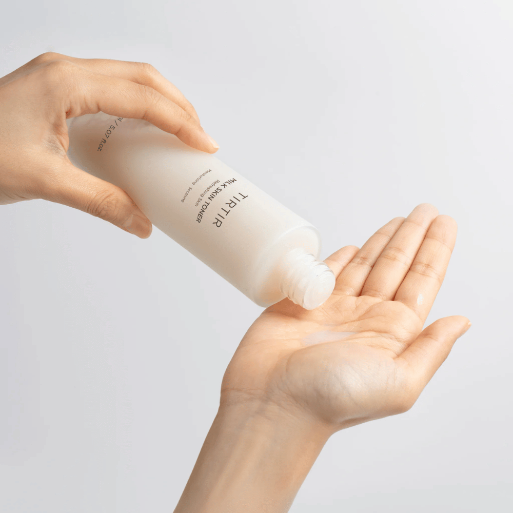 La imagen muestra a una persona vertiendo un líquido lechoso de una botella etiquetada "TIRTIR Milk Skin" en la palma de la mano. El producto parece ser un tónico facial o una loción hidratante.